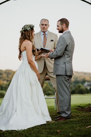 Outdoor wedding ceremony taking place under floral arch with scenic rolling hills in background.