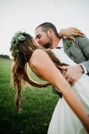 Romantic wedding couple shares an intimate dip kiss in a green field with floral crown details.