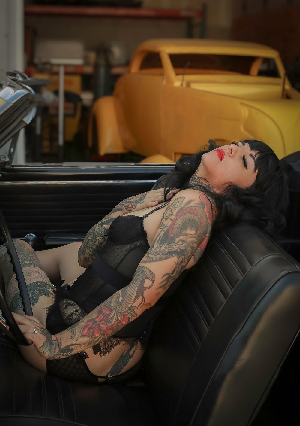 Reclining pinup model with tattoos lounges in black vintage convertible interior with classic cars visible.