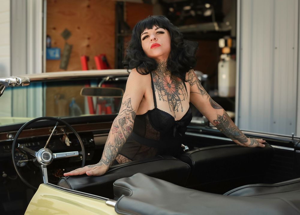 Person with tattoos and dark hair poses inside vintage car wearing black corset style top.