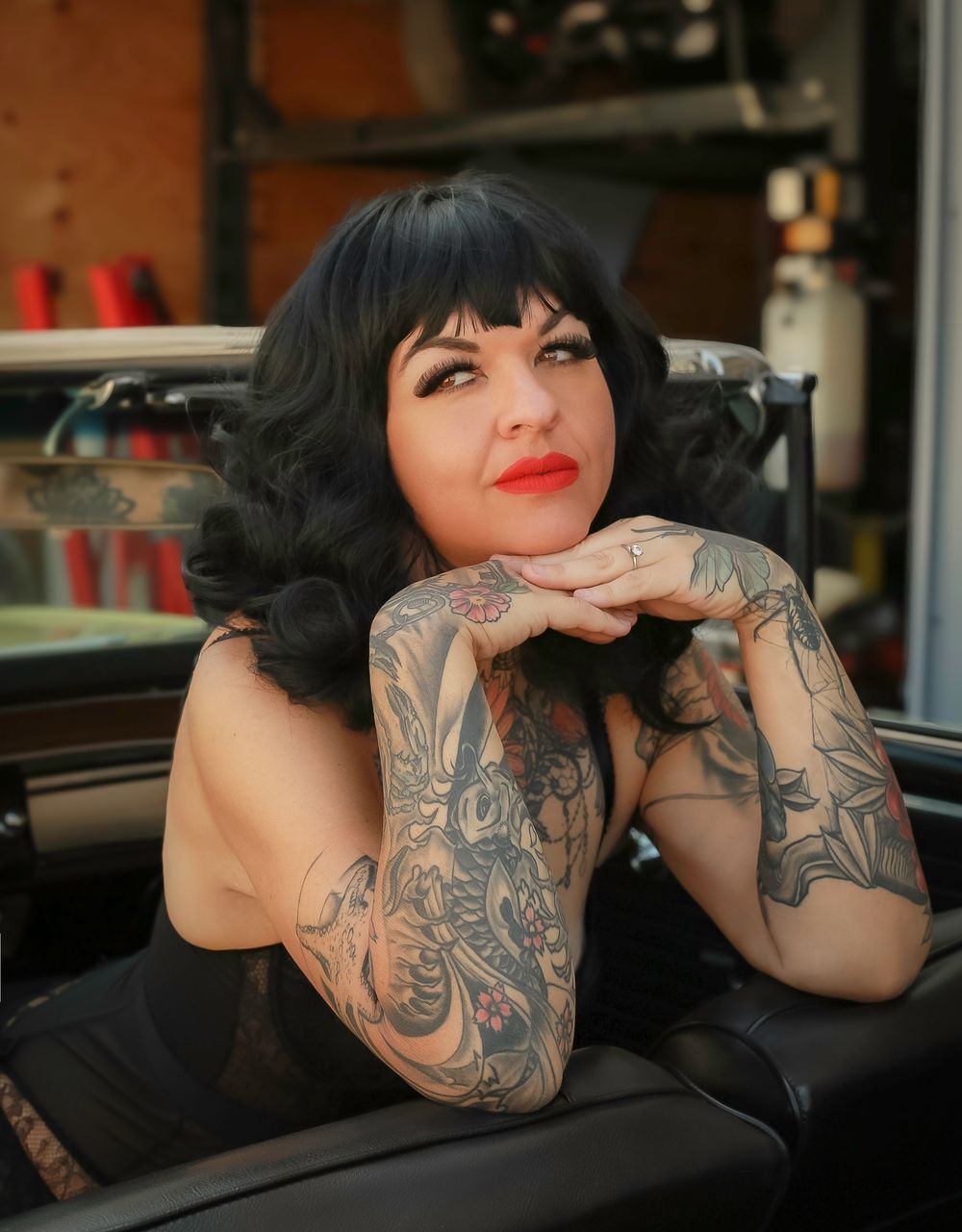 Retro styled model with dark curled hair and sleeve tattoos poses in classic car while resting on folded arms.