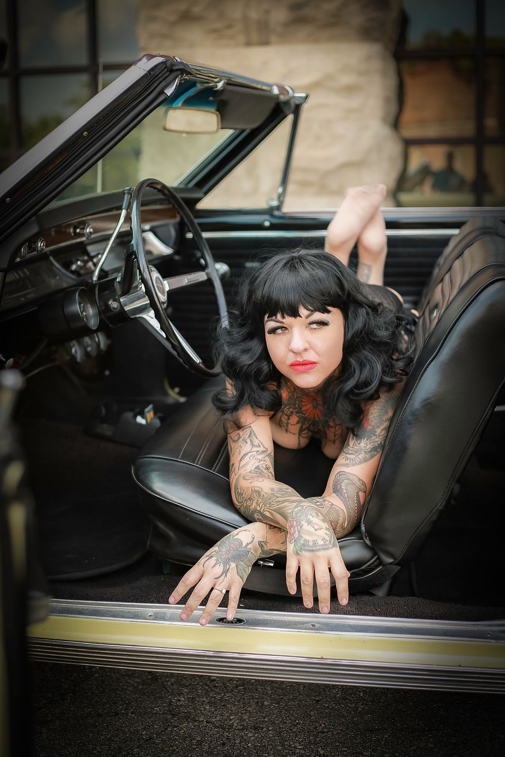 Vintage car interior photo featuring dark-haired model with tattoos in black outfit.