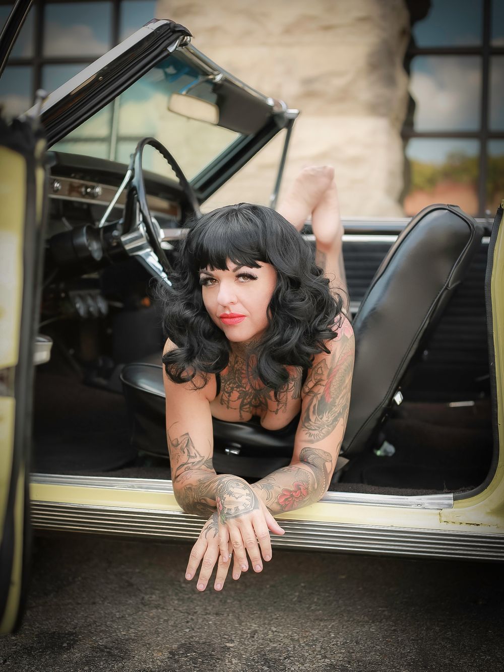 Retro pin-up style portrait in vintage car showing heavily tattooed model in black dress with dark curled hair.