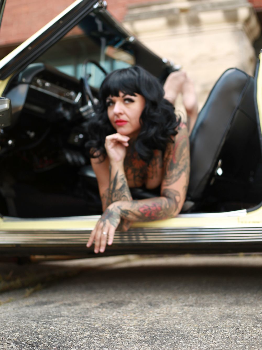 A vintage car portrait featuring a person with black hair, tattoos and a leather jacket leaning on the car door.