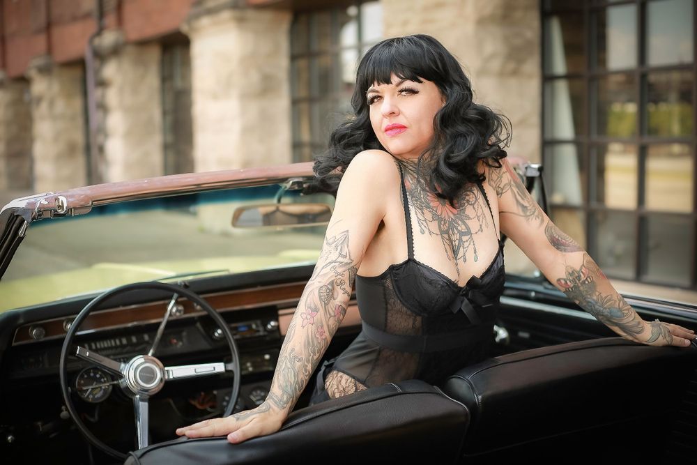 Glamorous vintage-style photo of tattooed model posing in classic convertible car with dark hair and makeup.