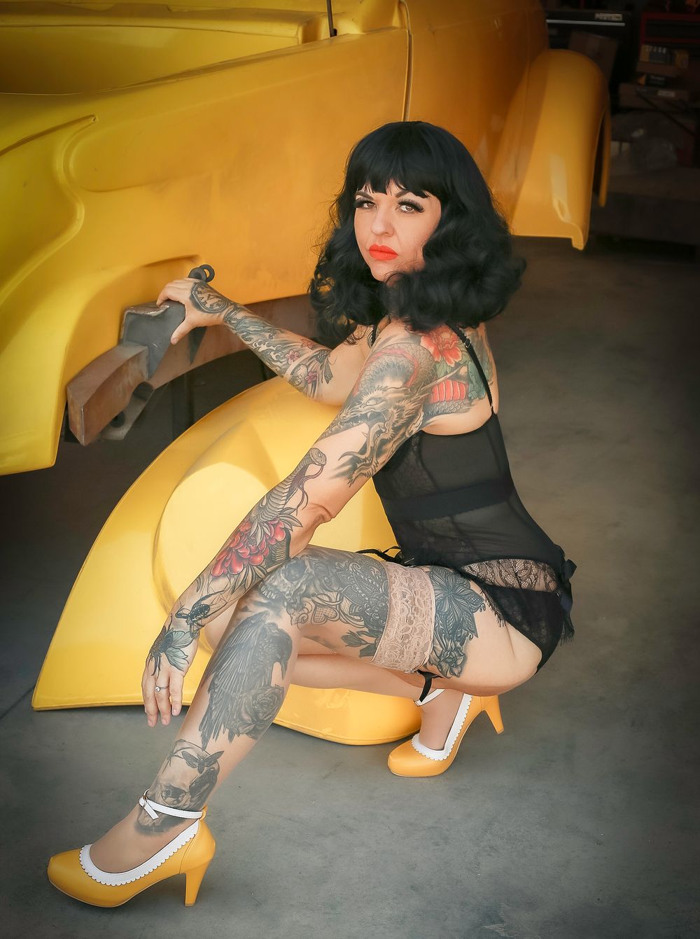 Tattooed model in vintage-style dress and heels poses against yellow background with retro aesthetic.
