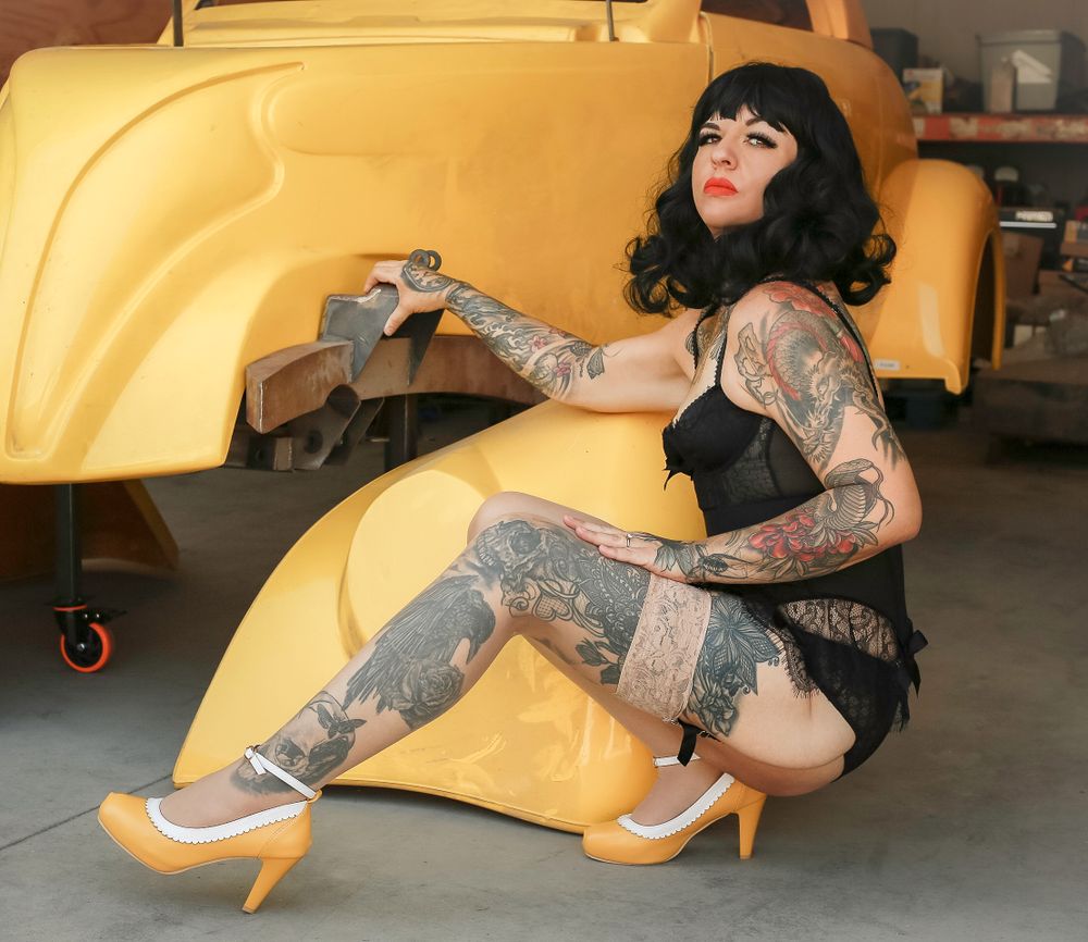 Gothic pin-up model in black dress with tattoos poses next to yellow vehicle in automotive garage setting.