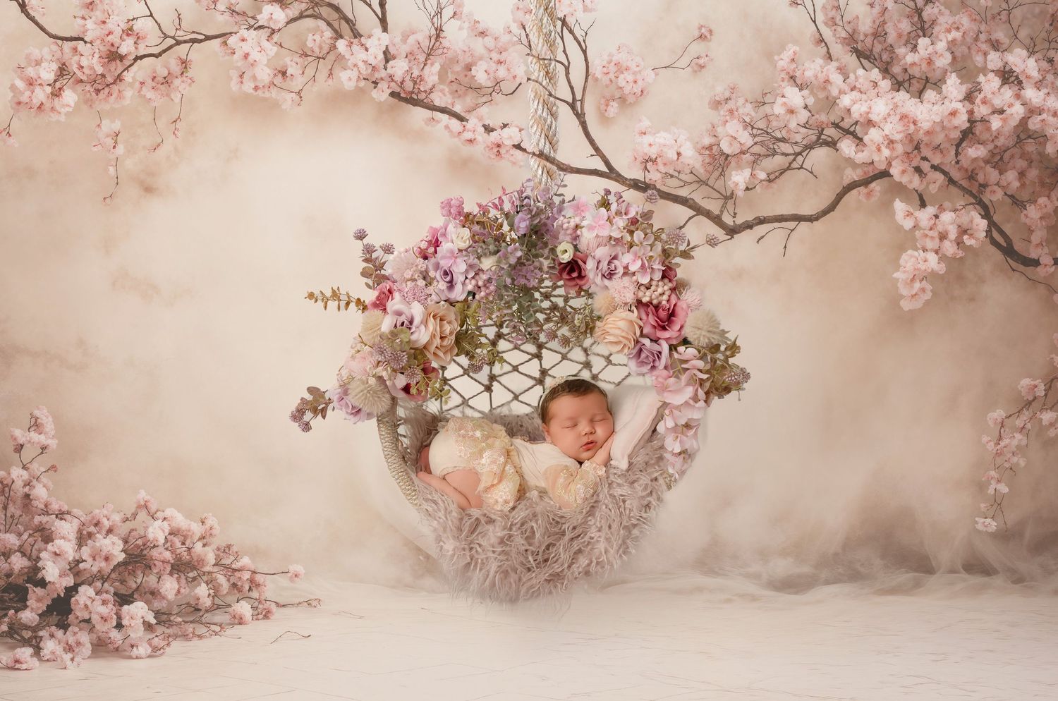 A sleeping newborn wrapped in pink fabric lies in a round basket surrounded by pink tulips and yellow flower buds on pink backdrop.