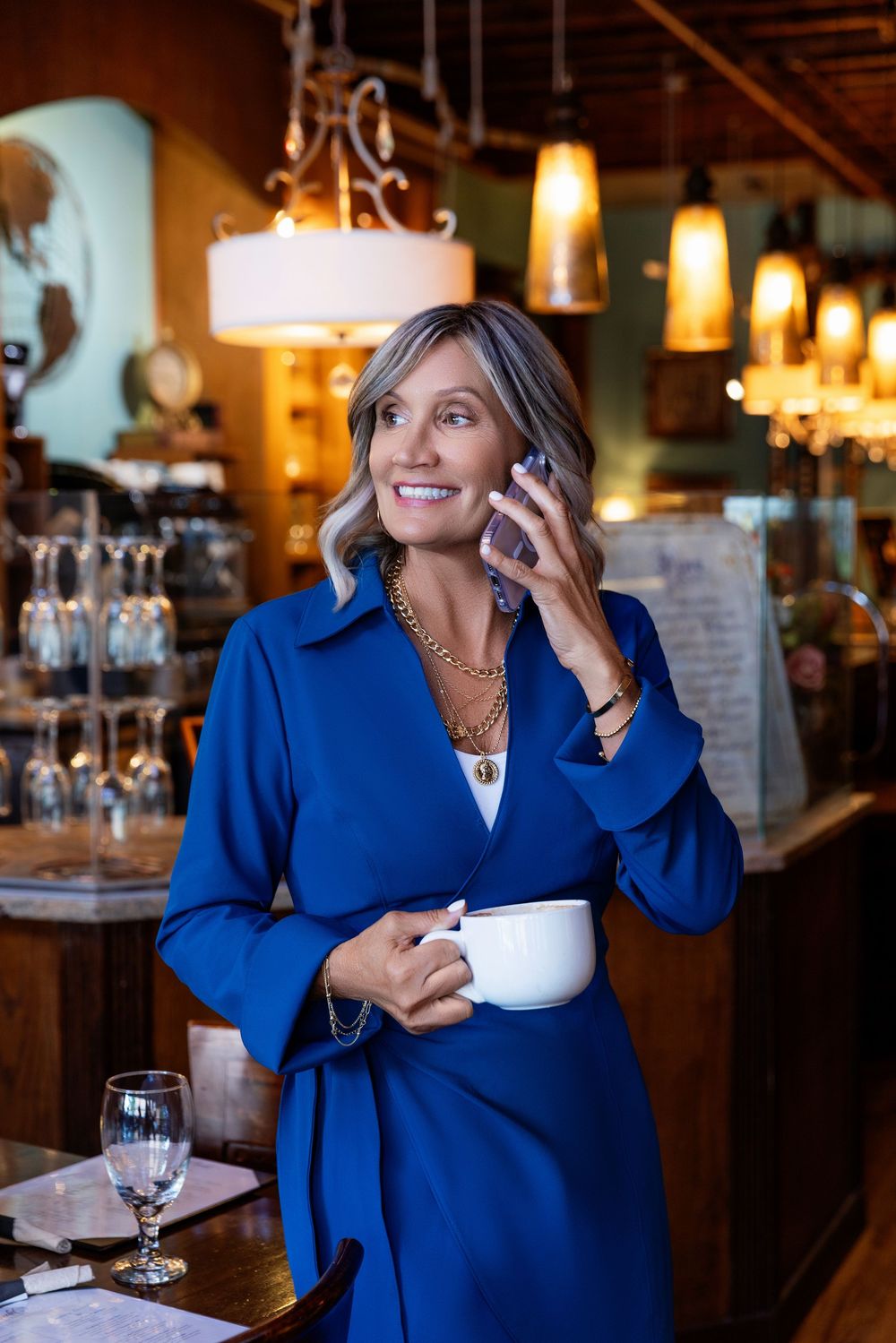 Woman in royal blue blazer sits at restaurant table with coffee cup and elegant lighting in warm interior setting.