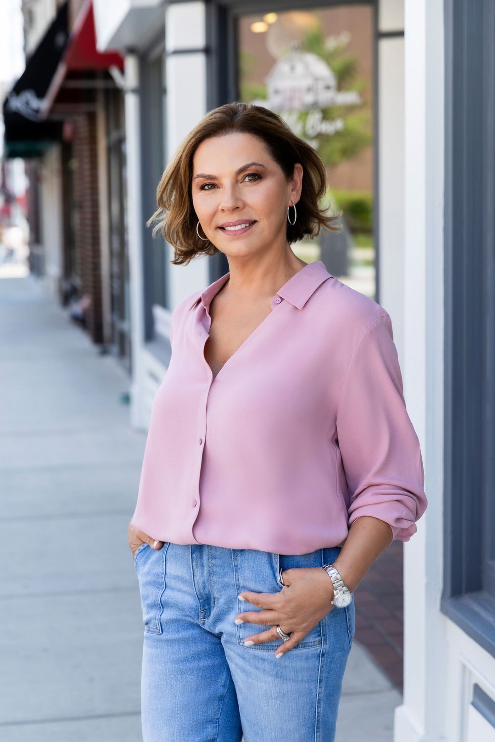 Woman in pink puff sleeve top and blue jeans poses in front of storefront windows.