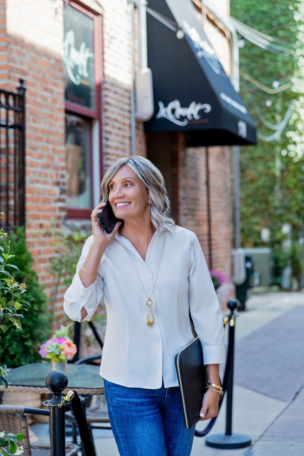 A person in a light colored top and jeans poses casually on a brick-lined street with storefronts and awnings.
