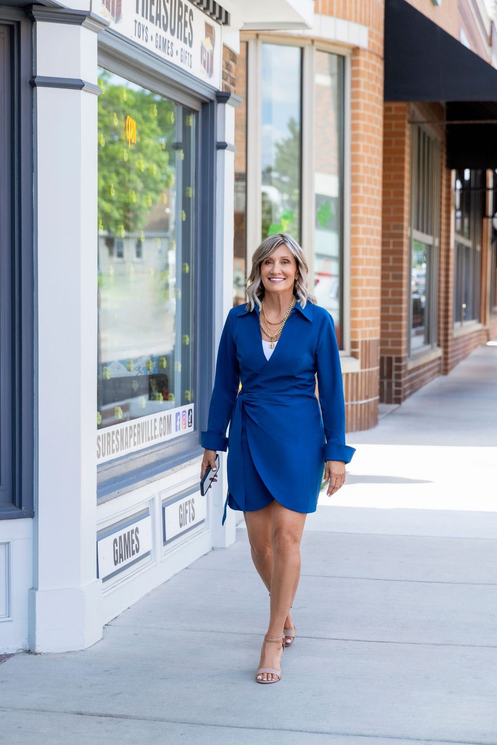Woman in royal blue blazer dress walks confidently along urban storefront sidewalk.