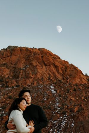Couple embracing romantically beneath a half moon above red rock mountain formations at dusk.