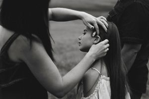 Black and white photo of parents gently touching their young daughters long hair outdoors.
