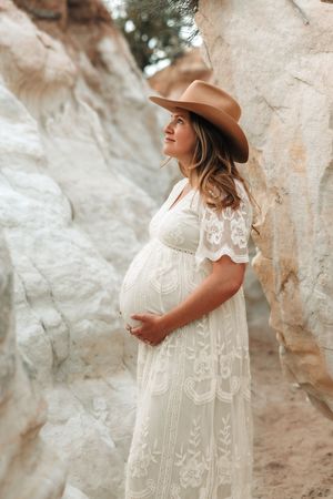Pregnant woman in white lace dress and brown cowboy hat posing gracefully among rocky formations.