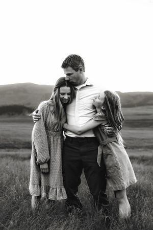 Black and white photo of a father embracing two daughters in an open field with rolling hills.