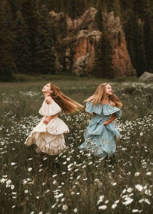 Two girls in flowing white and blue dresses twirl joyfully in a wildflower meadow near red rock cliffs.