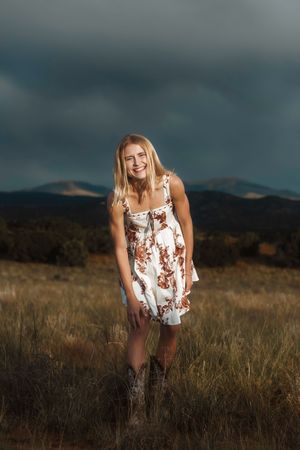 Colorado senior in floral sundress and cowboy boots standing in field with stormy sky and mountains