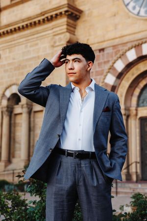 Young man in gray suit with red pocket square poses confidently before historic stone building.