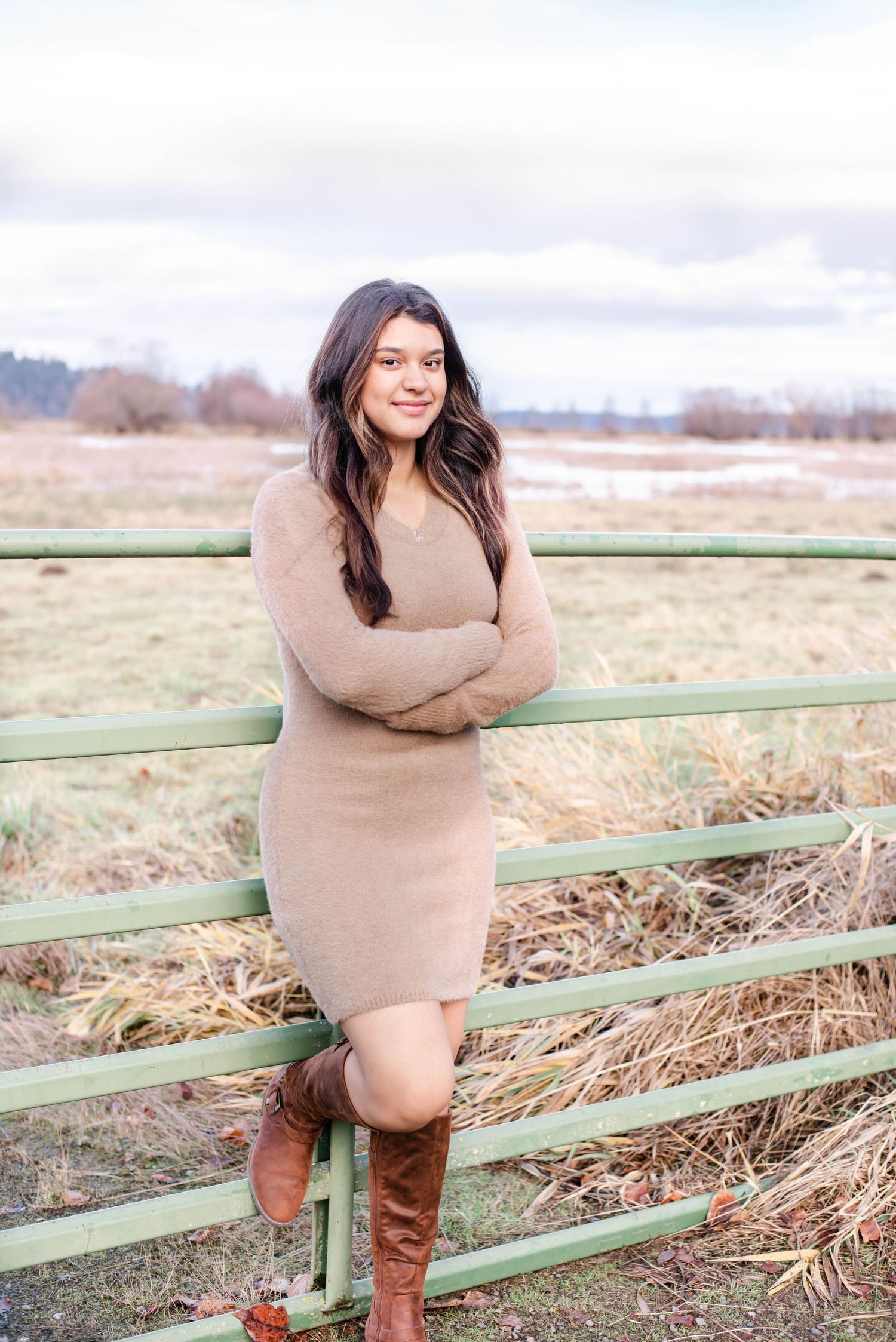 Person in beige sweater poses by rustic green fence in a winter field setting.