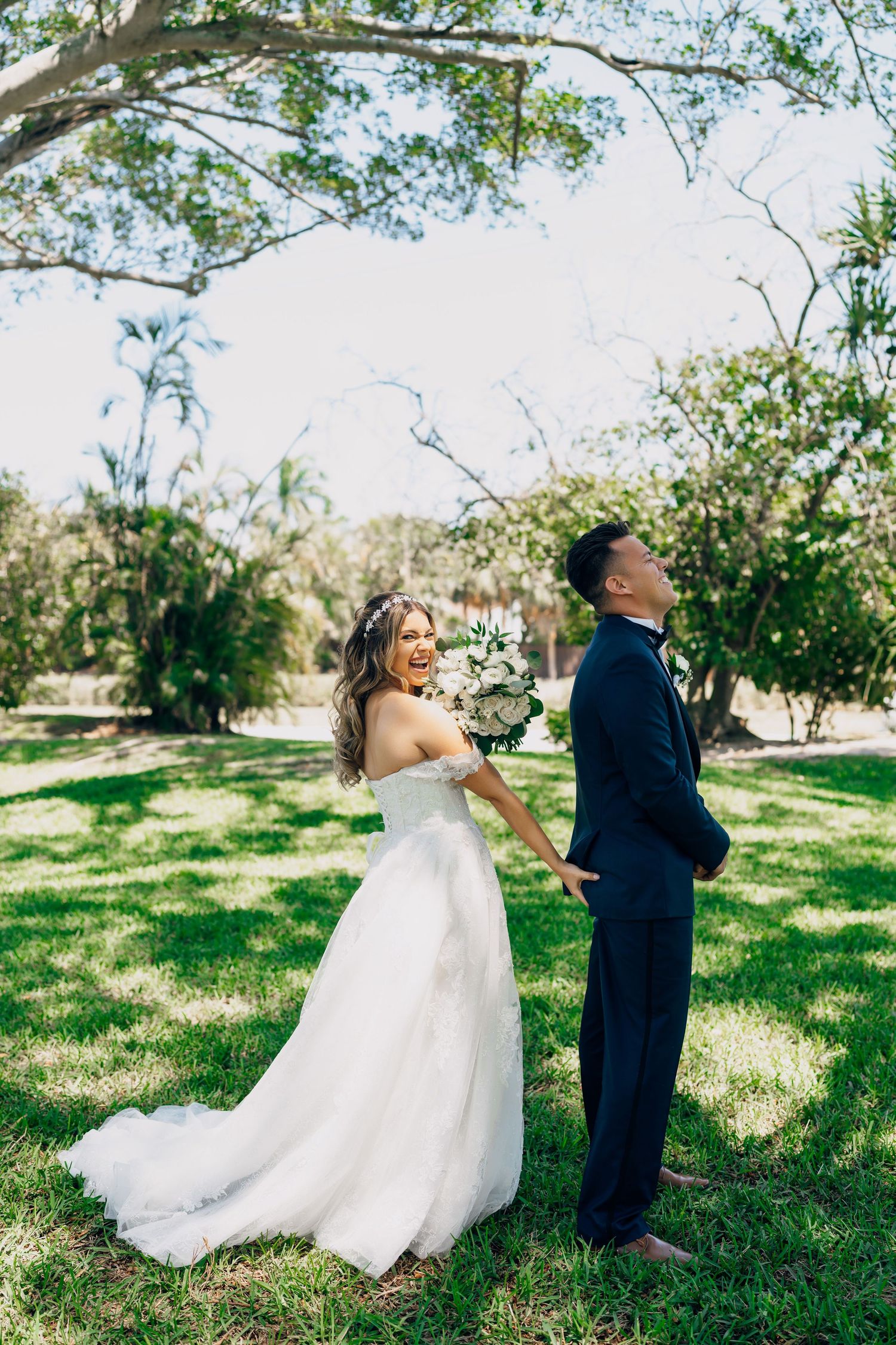 A bride in a flowing white gown shares a playful first look moment with her groom in a garden setting.