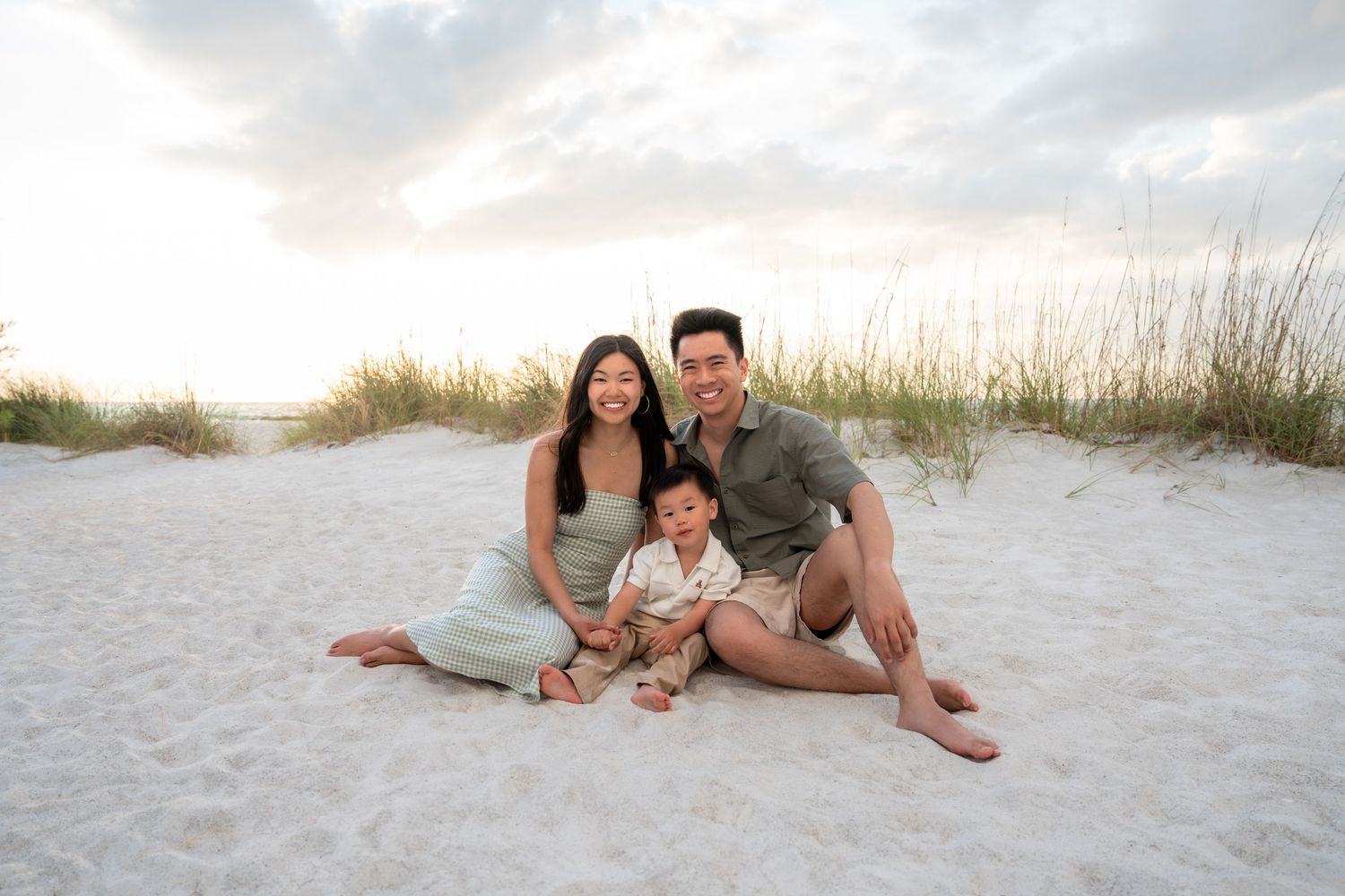 Happy Asian family sitting on white sand beach at sunset, parents smiling with toddler son.