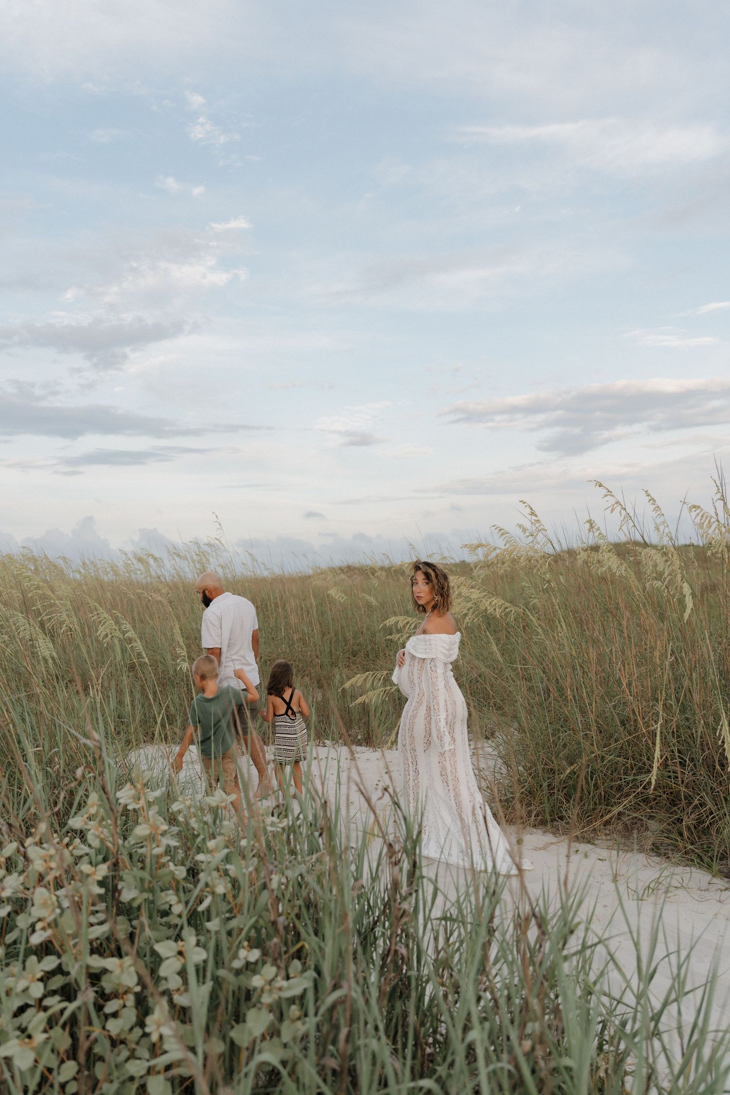 A couple in white clothing walks on a beach while seagulls fly overhead at sunset.