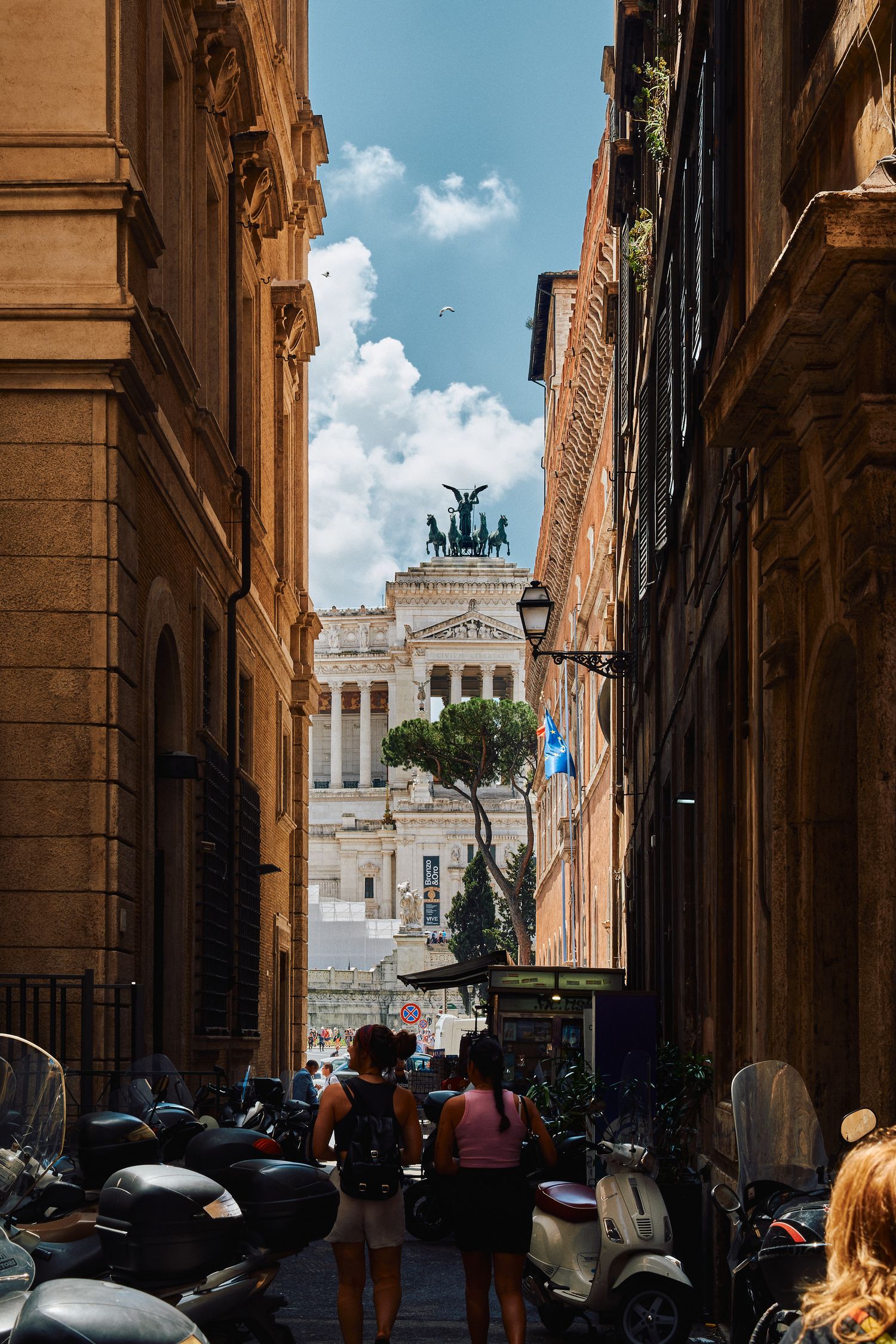 View of Vittorio Emanuele II Monument through narrow Roman alley filled with parked scooters.
