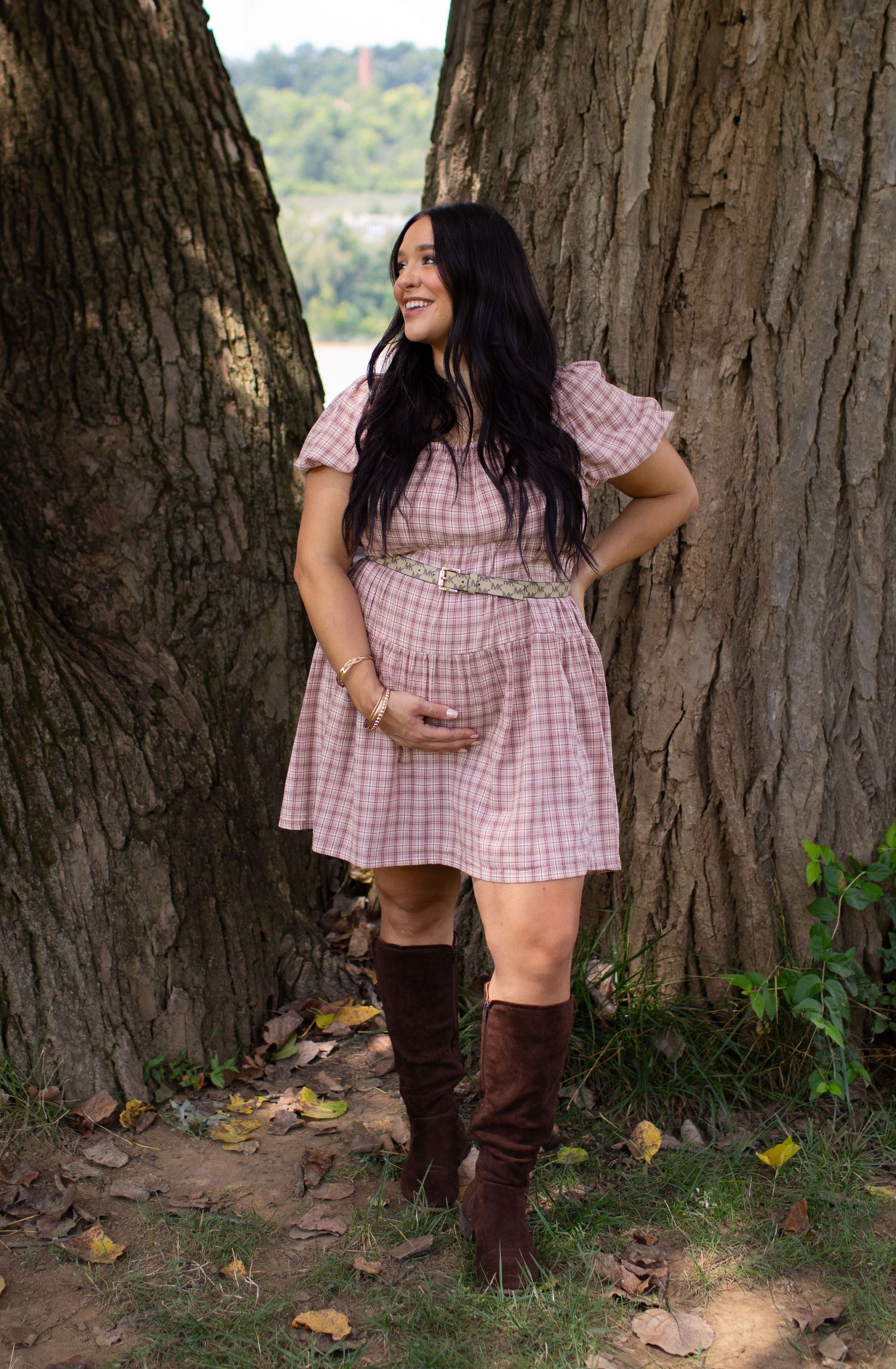 Pregnant woman in pink plaid dress and brown boots poses between two large trees.
