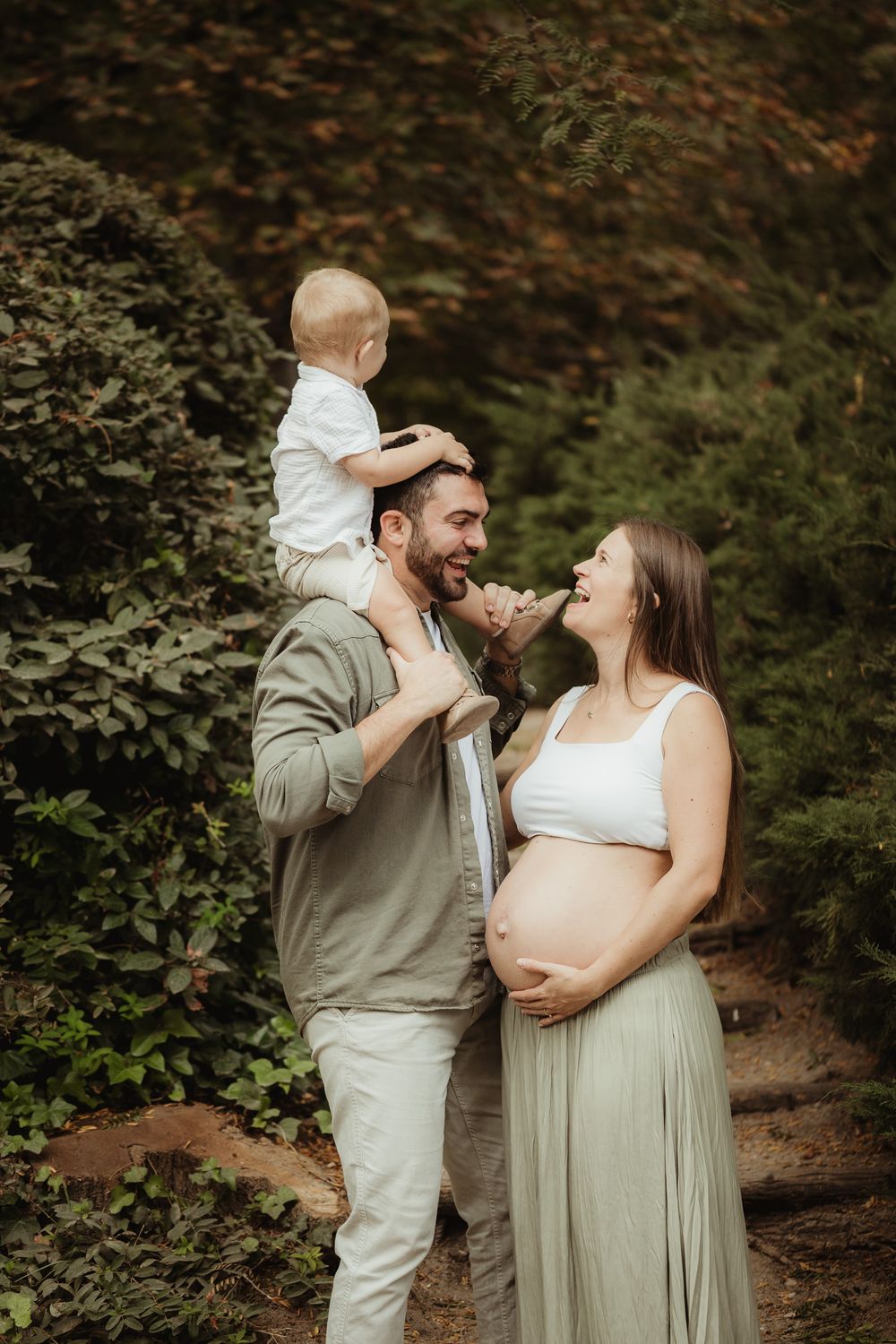 Pregnant woman and man with toddler on shoulders share a joyful moment outdoors surrounded by lush green foliage.