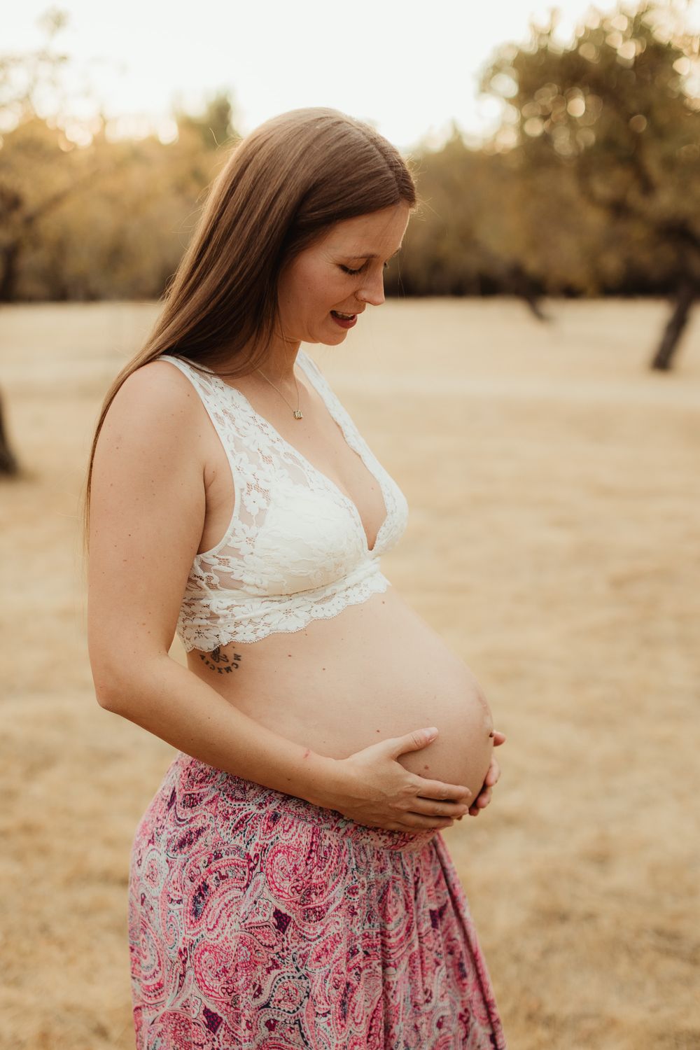 Pregnant woman in white lace bralette and pink paisley skirt cradling her baby bump outdoors at golden hour.