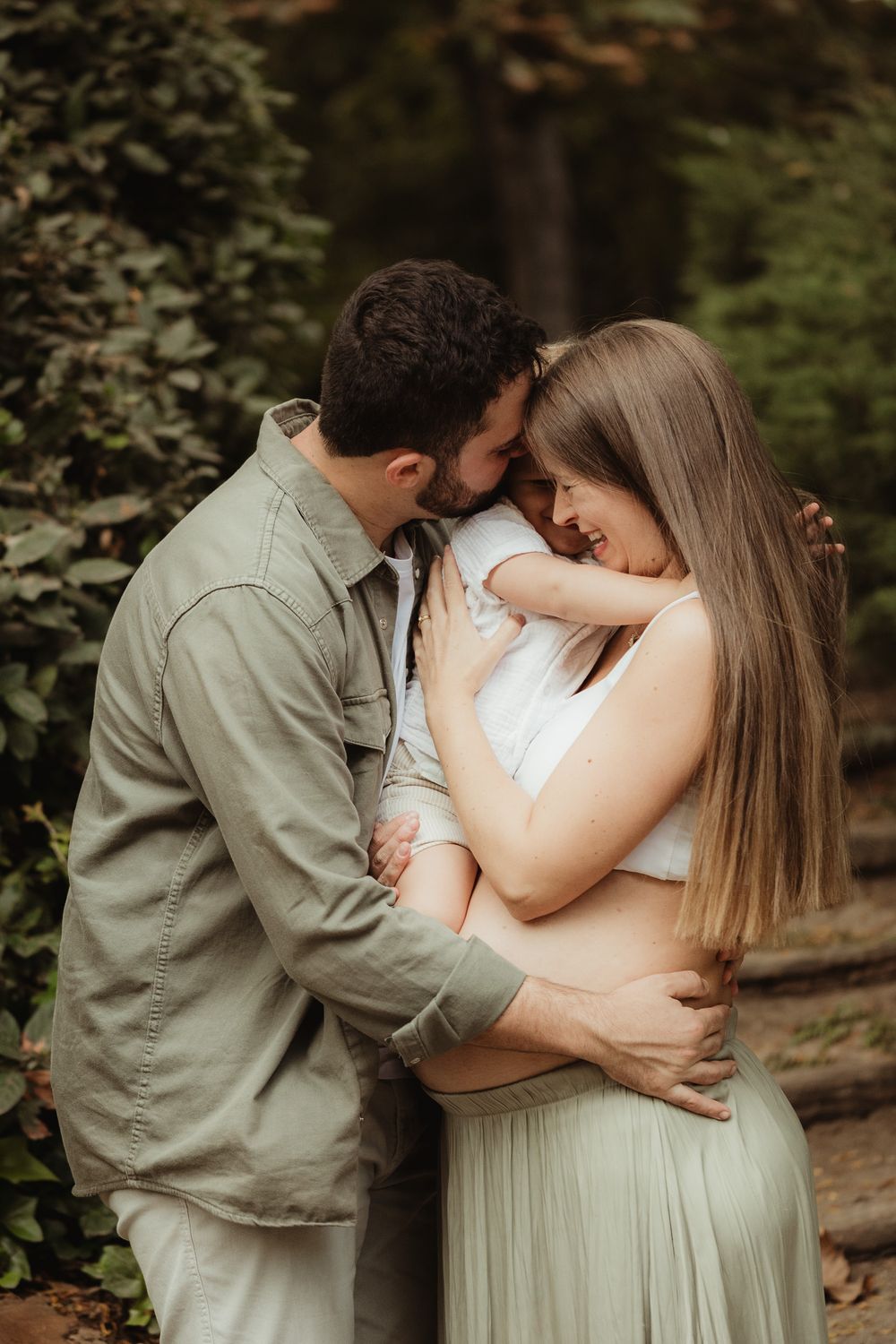 Expecting couple embracing in forest setting, man kissing womans forehead, pregnant belly visible, wearing neutral earth tones.