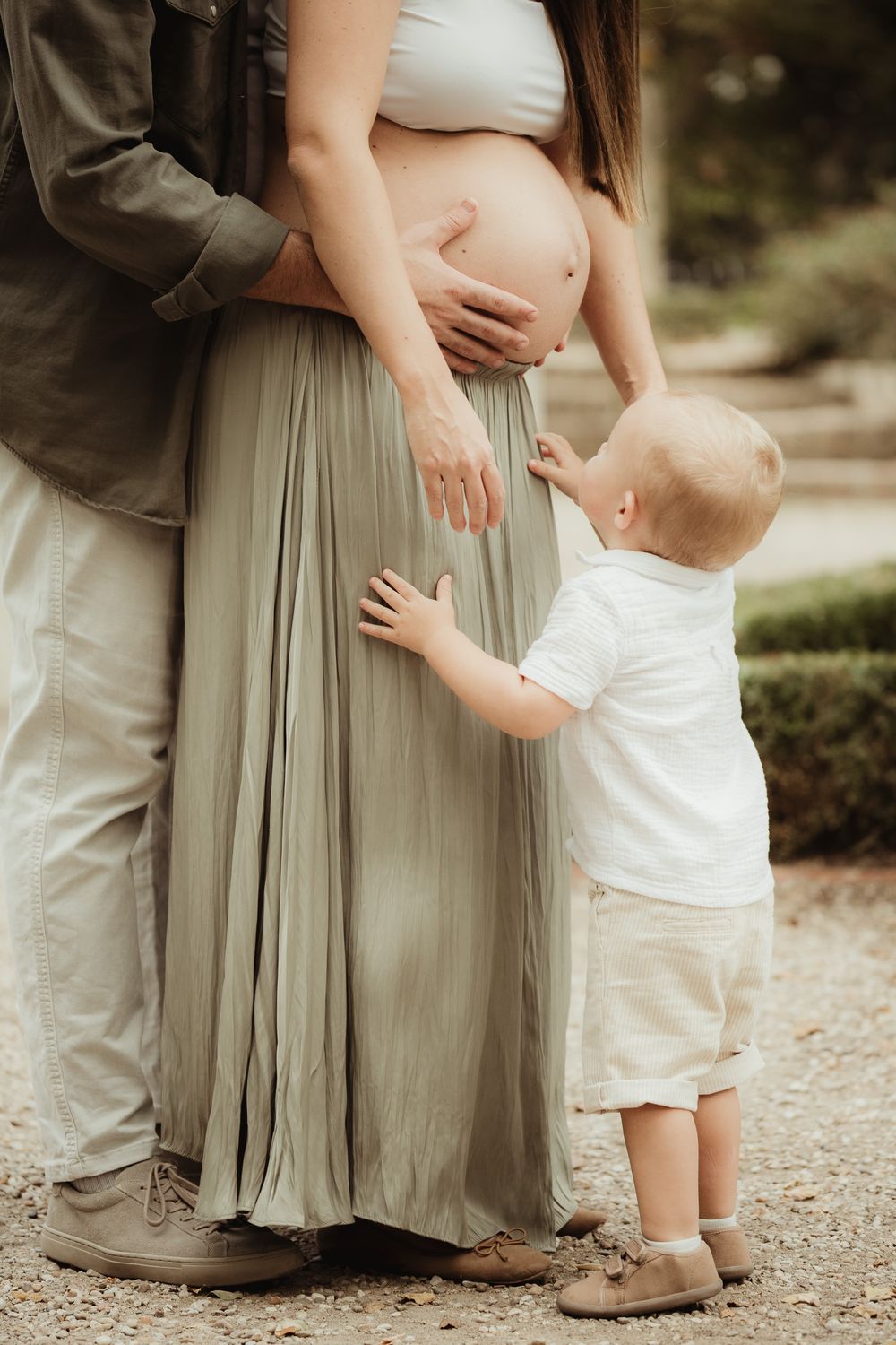 Pregnant woman in sage green skirt with toddler touching her belly, standing beside partner outdoors.