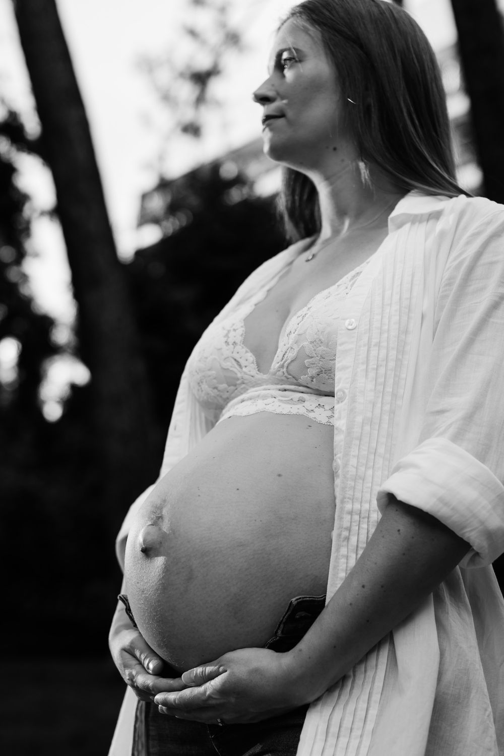 Black and white portrait of a pregnant woman outdoors, cradling her bare baby bump, wearing a white lace bra and open shirt.