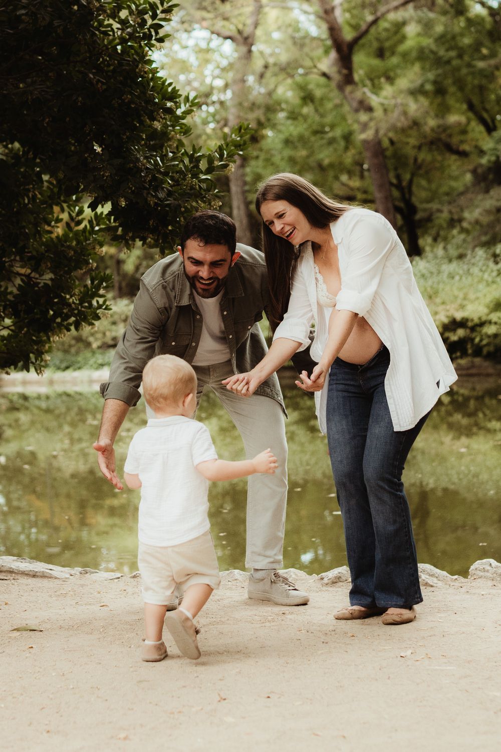 Joyful family with pregnant mother, father, and toddler playing outdoors near a pond surrounded by lush green trees.