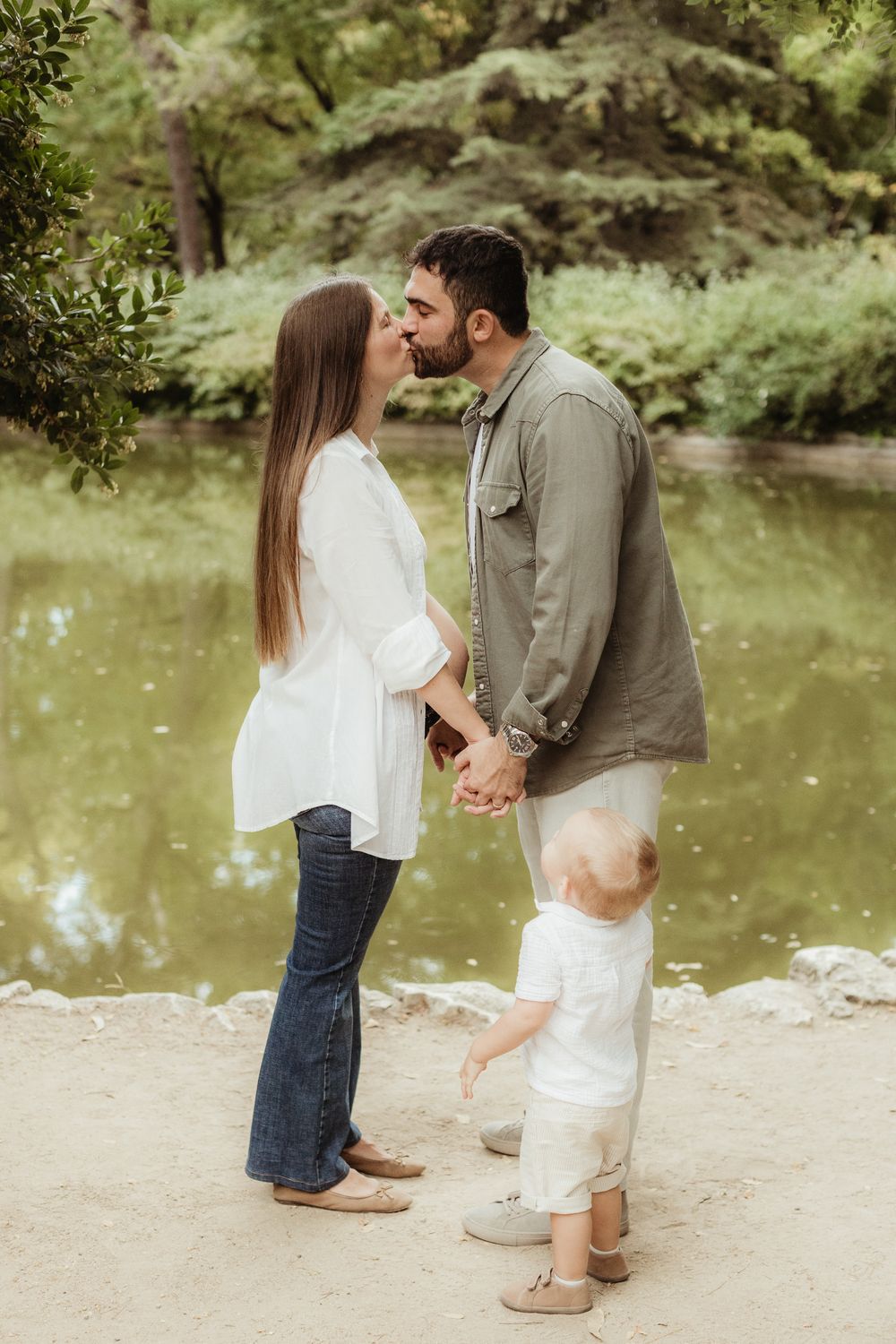 A couple kisses by a pond in a park while holding hands with their toddler standing between them.