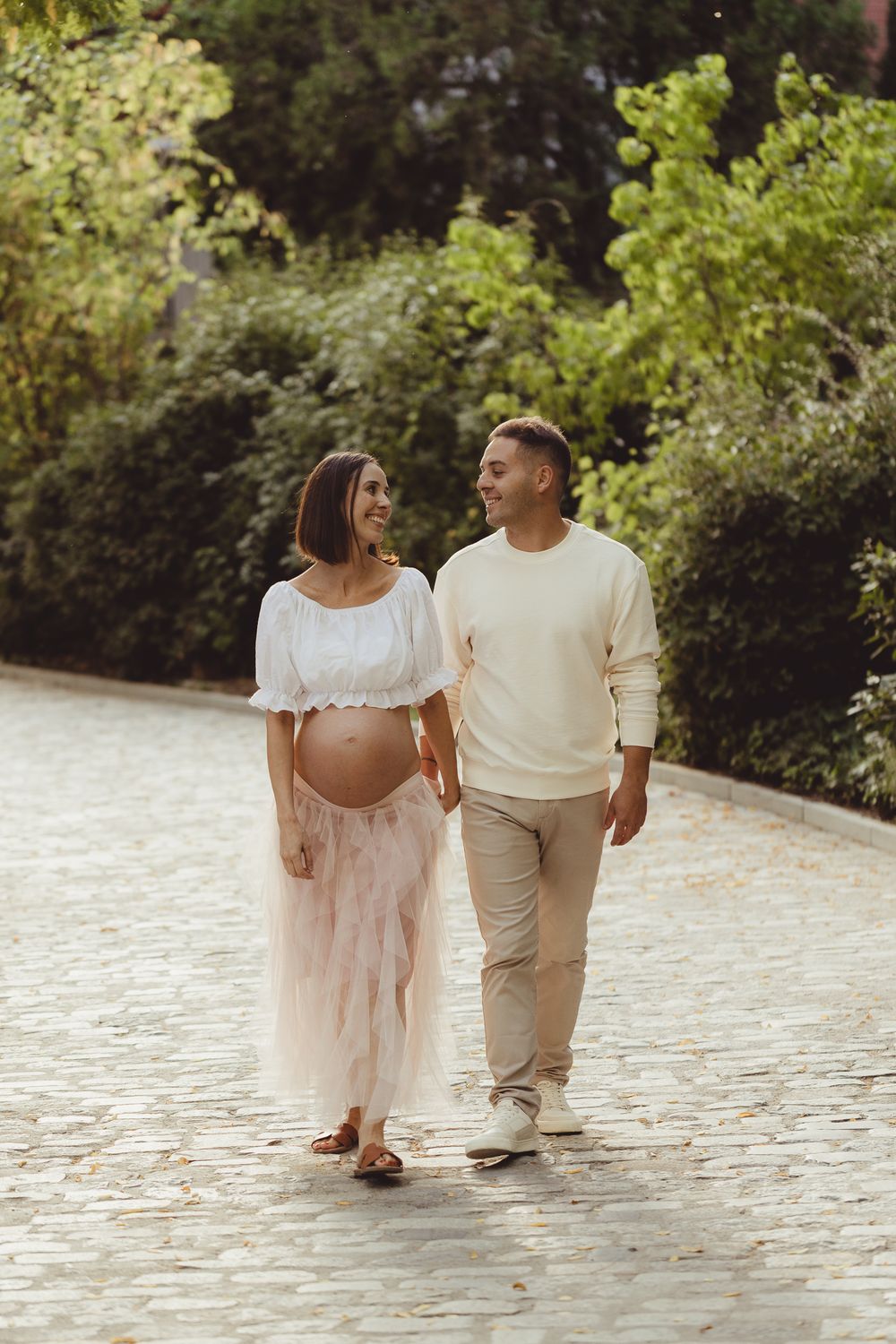 A couple in light colored outfits walking together on a stone path surrounded by lush greenery.