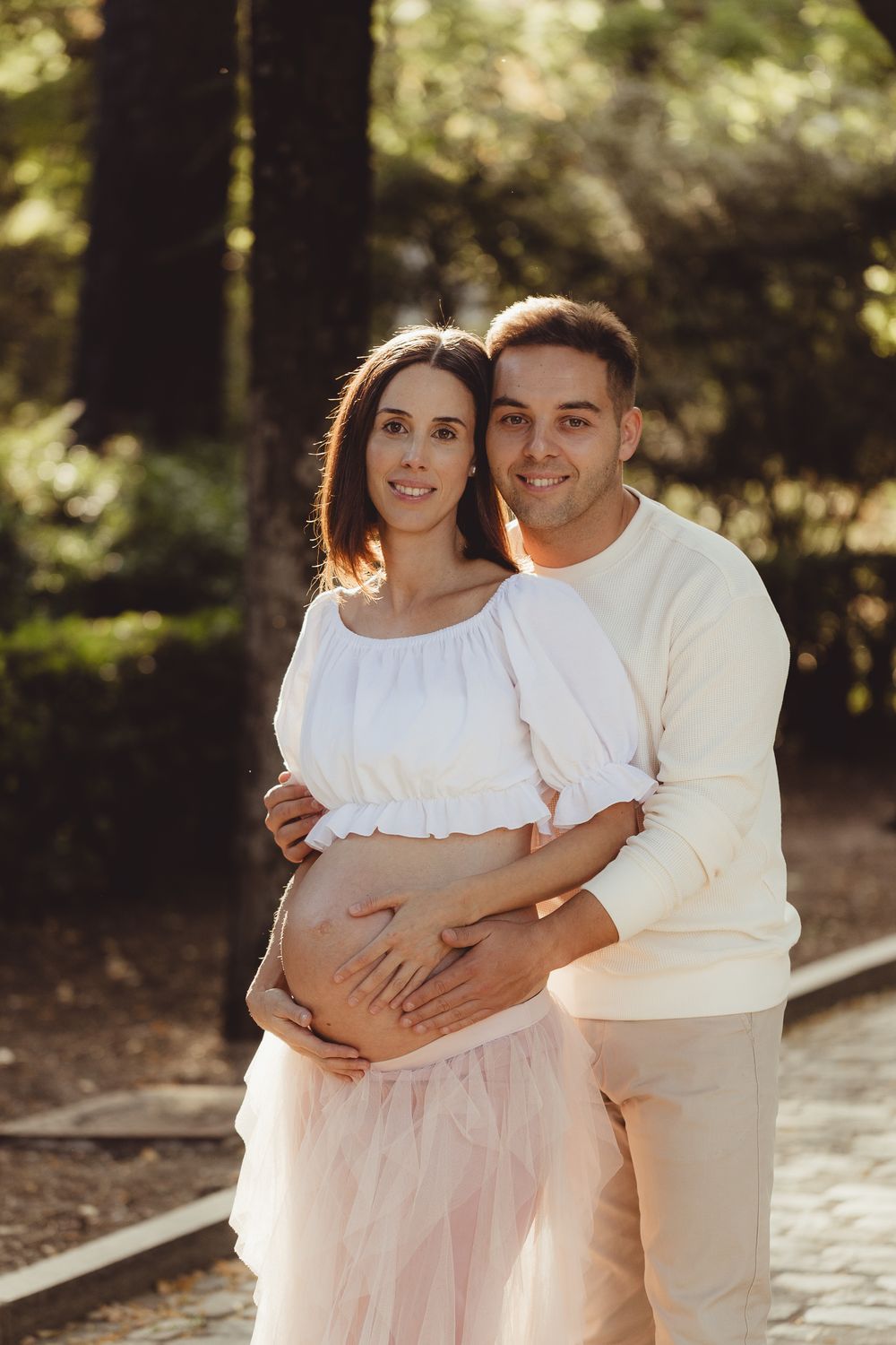 Couple in white outfits embraces during outdoor maternity photoshoot with warm sunlit backdrop.