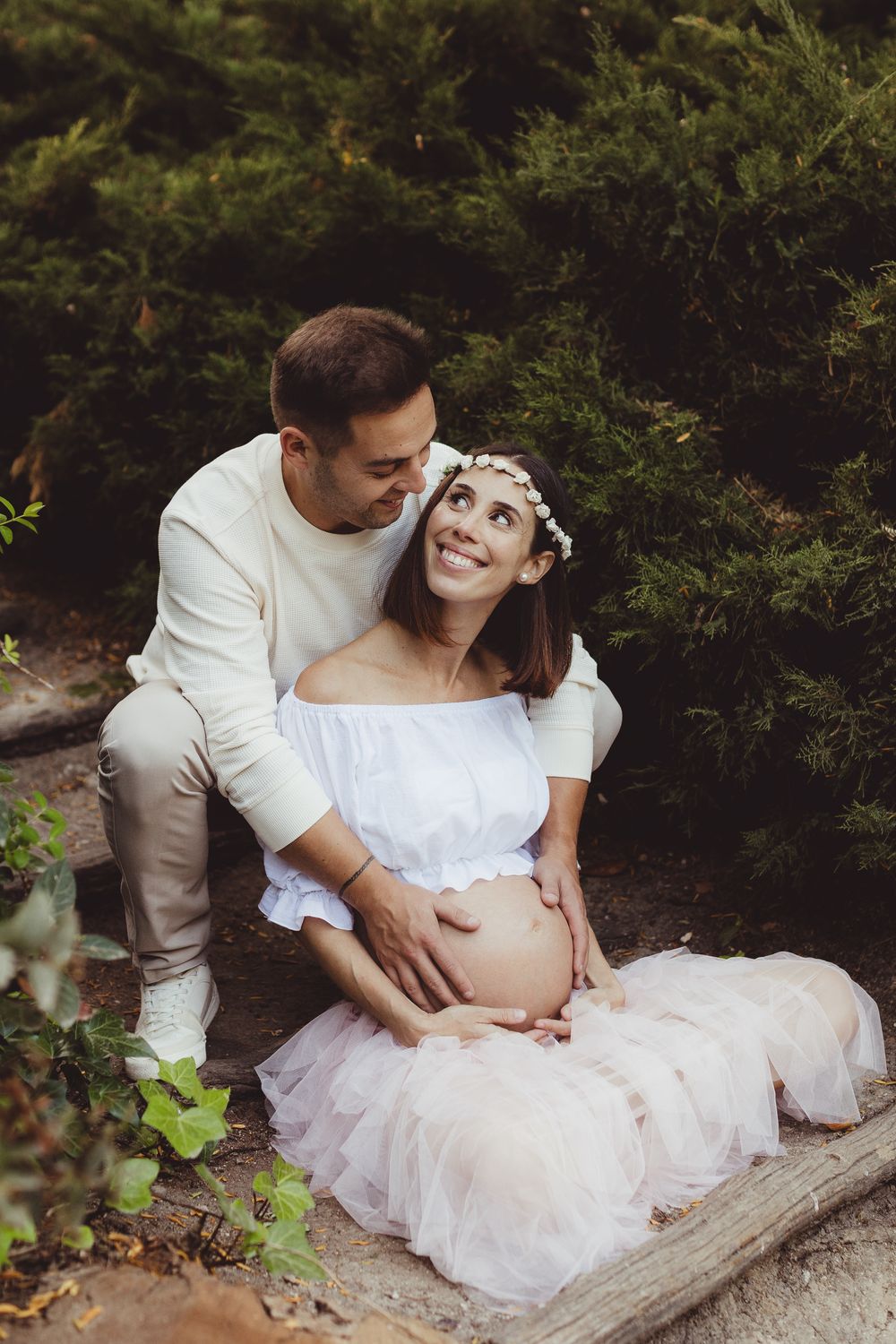 A romantic maternity photoshoot in nature shows a couple sitting on a log surrounded by lush greenery.