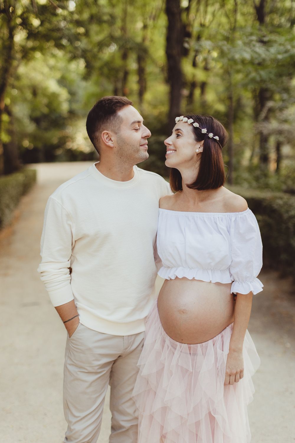 A romantic maternity photoshoot in a wooded setting with a couple wearing white and pink outfits.