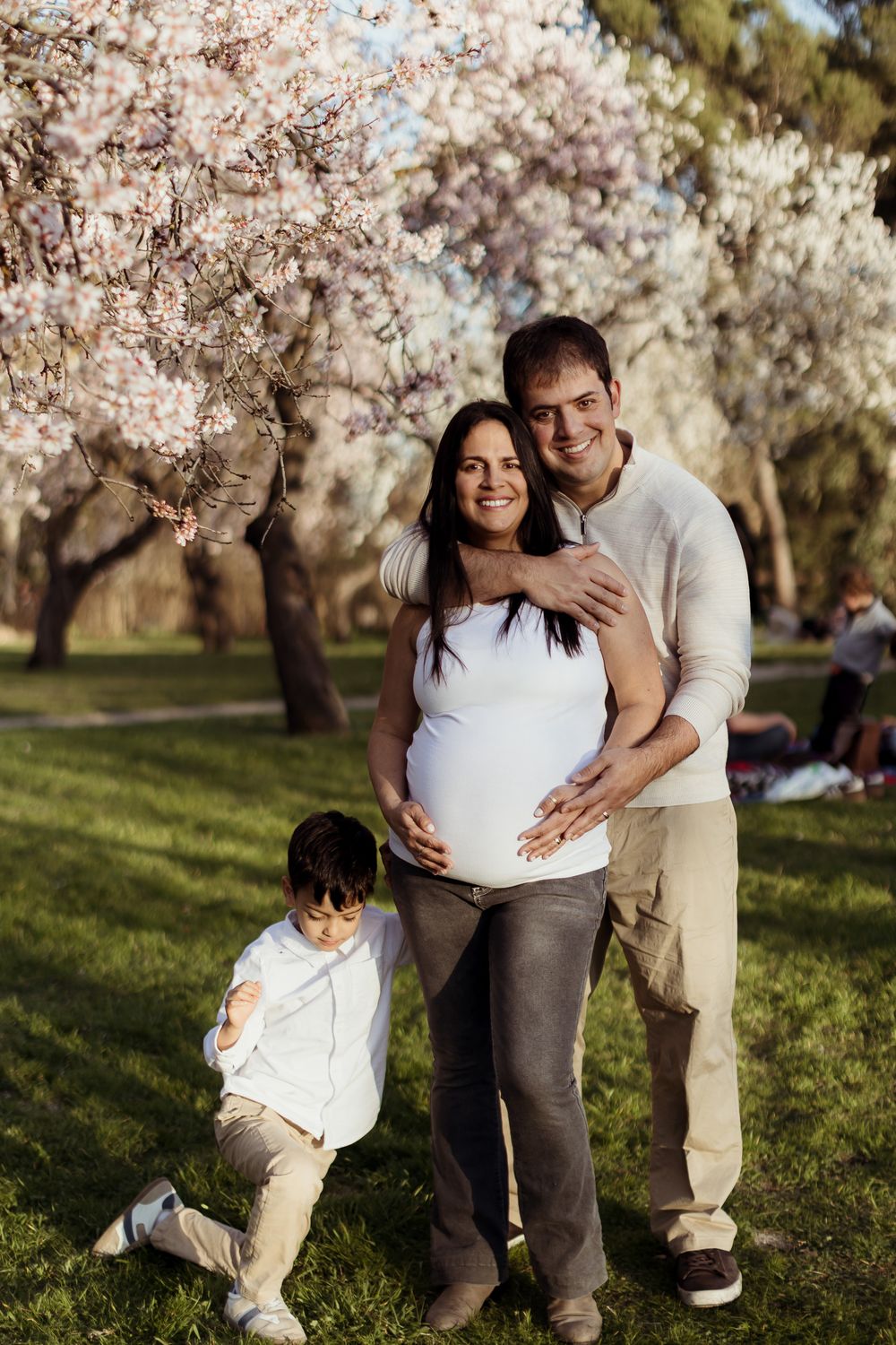 Pregnant woman with husband and child posing in blooming spring park with white cherry blossom trees.
