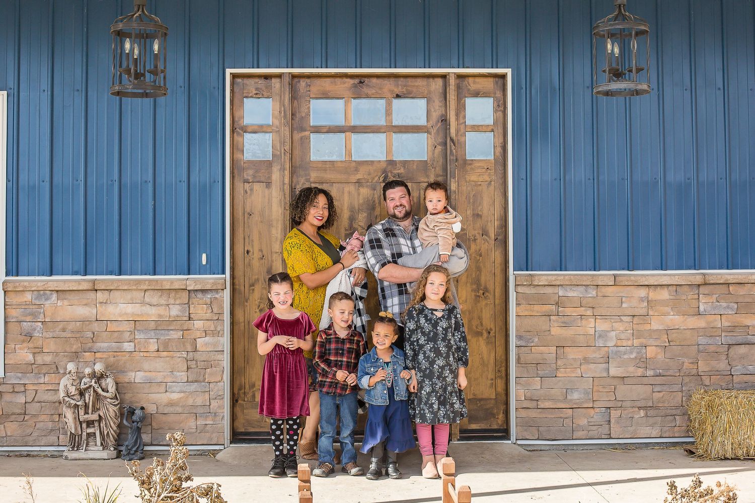 Happy family of seven posing together in front of a rustic blue barn with stone accents and wooden doors.