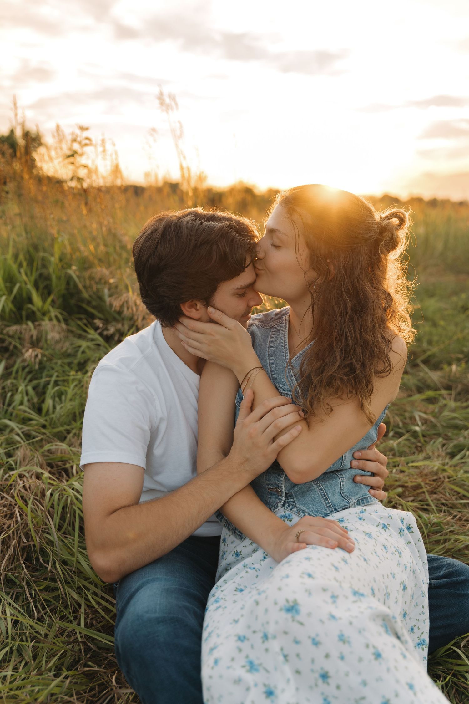 A tender sunset kiss shared between a couple sitting in a golden field of tall grass.