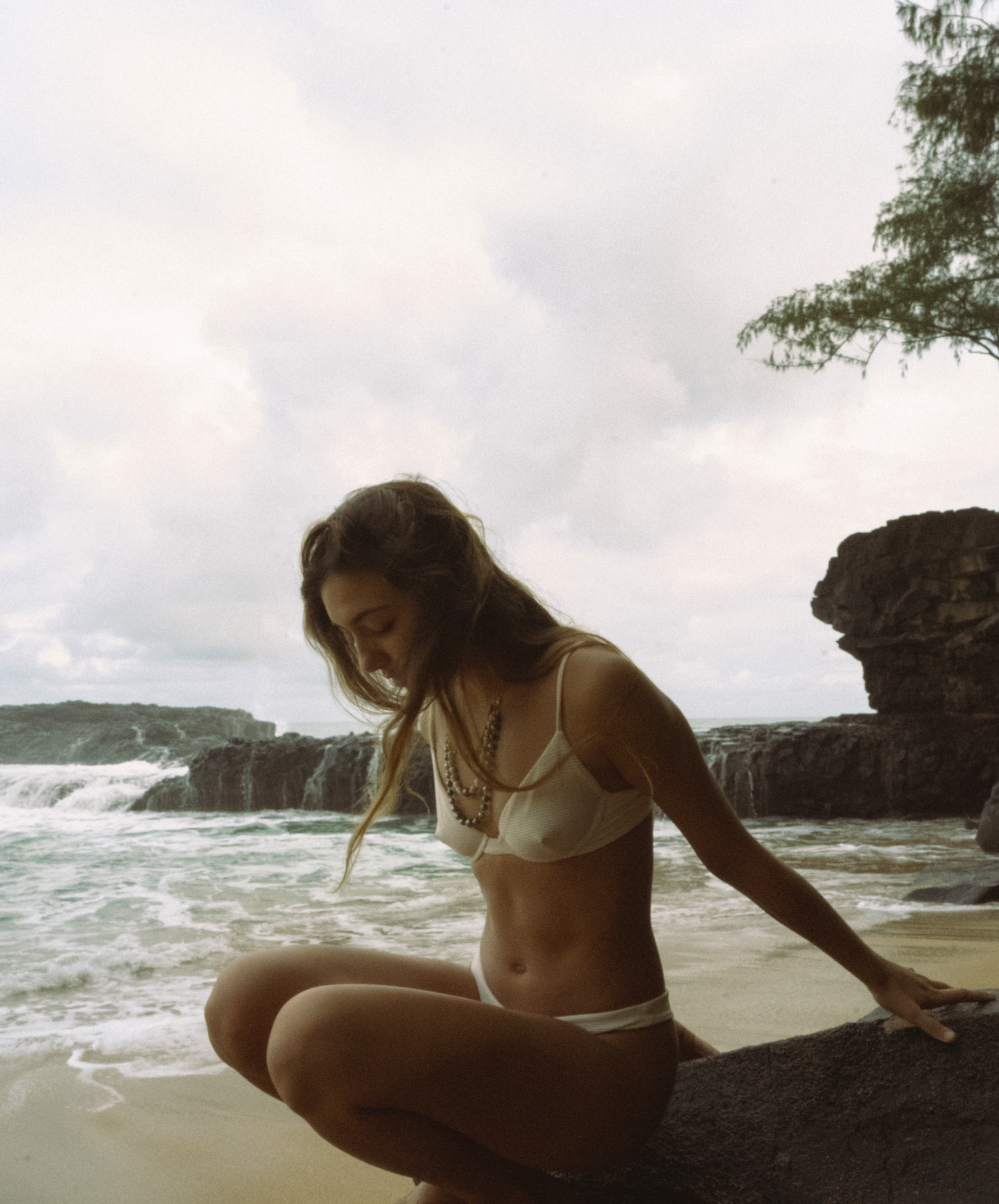 a girl sitting on a rock looking in the sand with water and waves surrounding her at tourist lumahai kauai