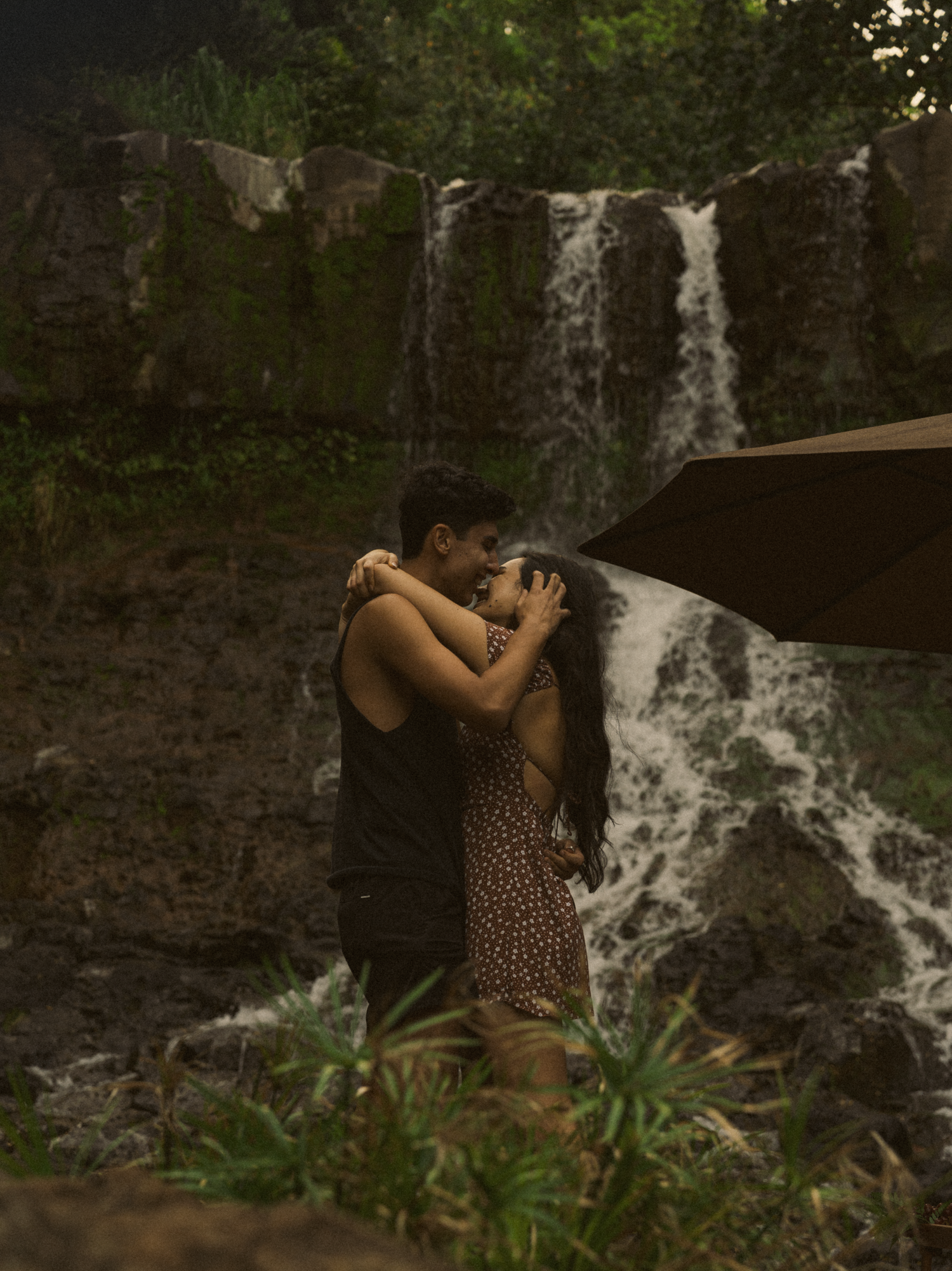 Couple embracing in front of a cascading waterfall in a lush forest setting.