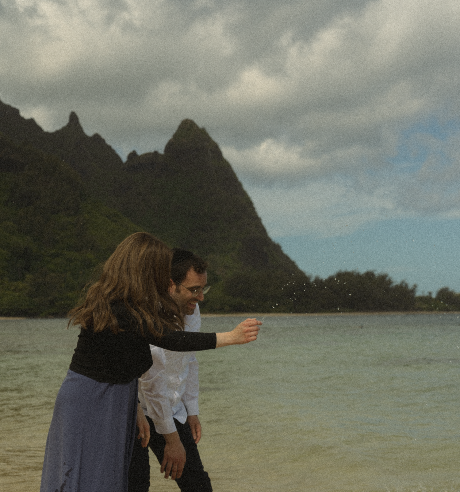 Two people splash and play in tropical waters with dramatic mountain peaks in the background.