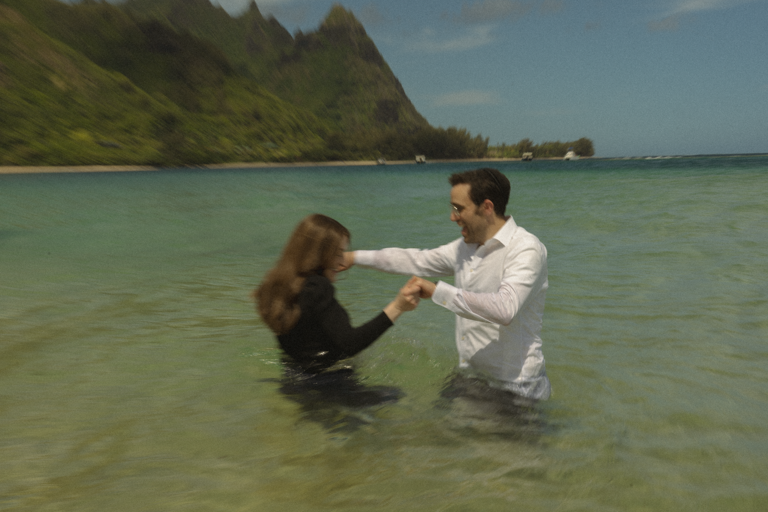Couple playing in turquoise ocean waters with lush mountainous coastline.