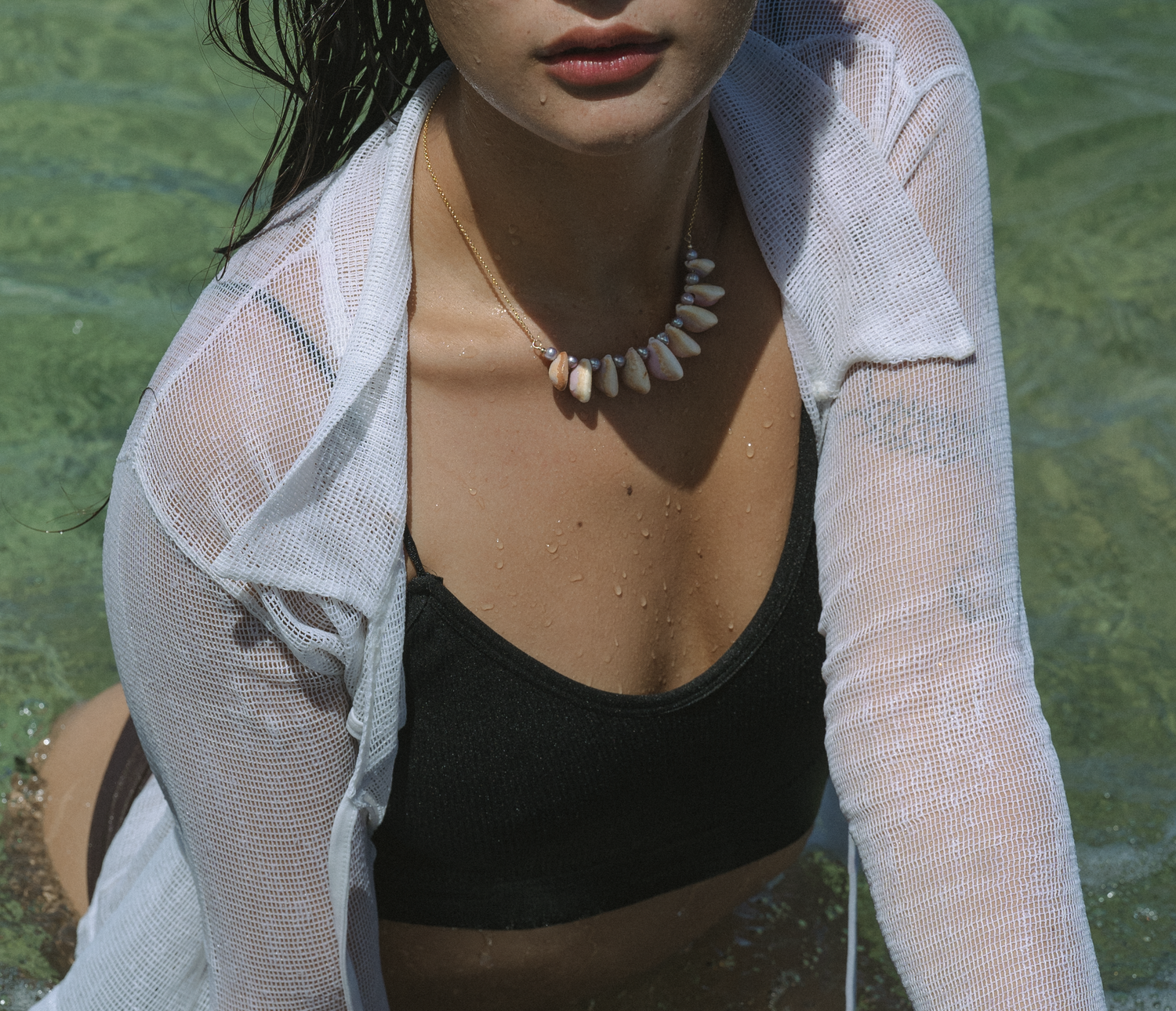 Woman in white sheer cover-up and black swimwear with pearl necklace by water.