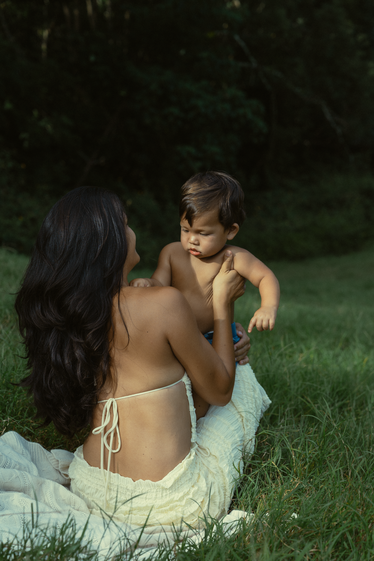 A tender moment between parent and child laying in green grass wearing white clothing at sunset.