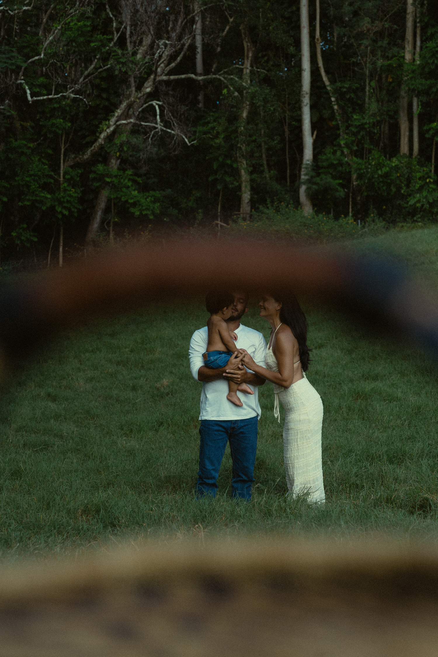 A romantic couple embraces in a serene outdoor setting with lush green trees in the background during golden hour.